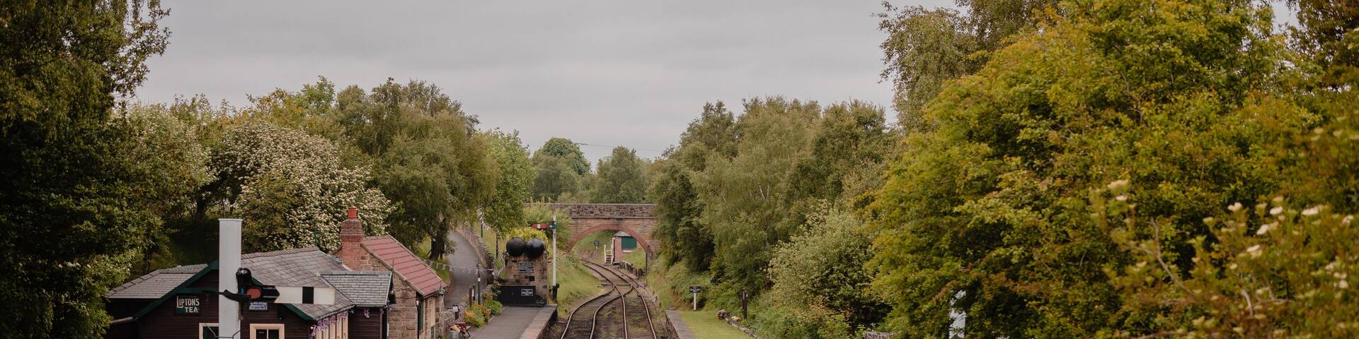 A railway station with tracks, benches, a small building, and a bridge in the background.
