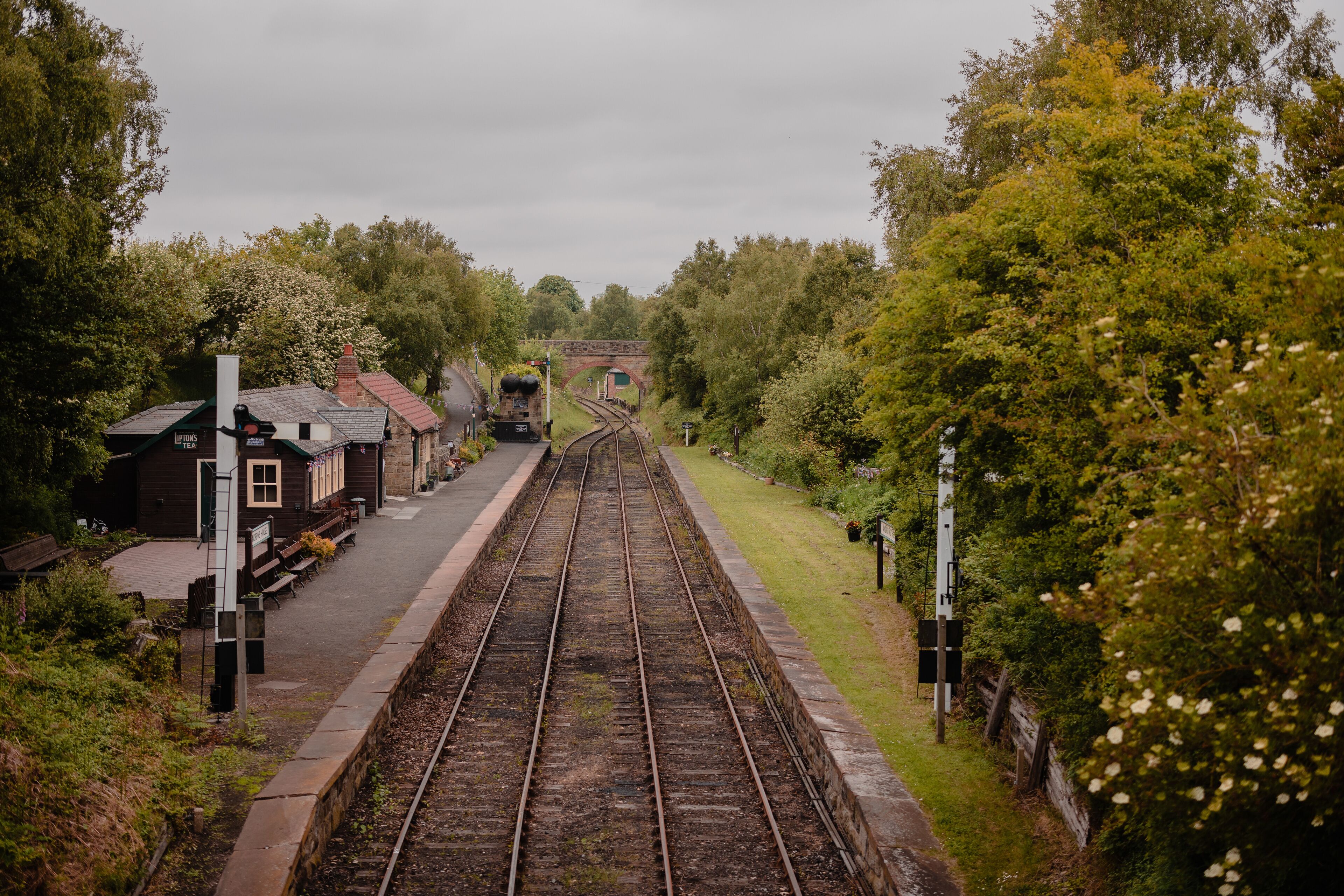 A railway station with tracks, benches, a small building, and a bridge in the background.