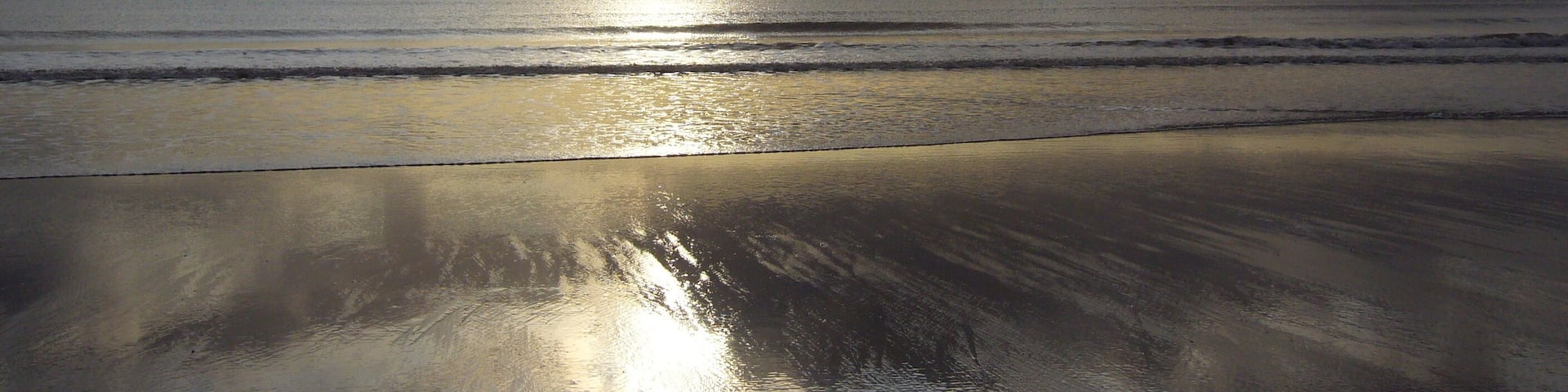A beach with wet sand reflecting sunlight and waves approaching the shore.