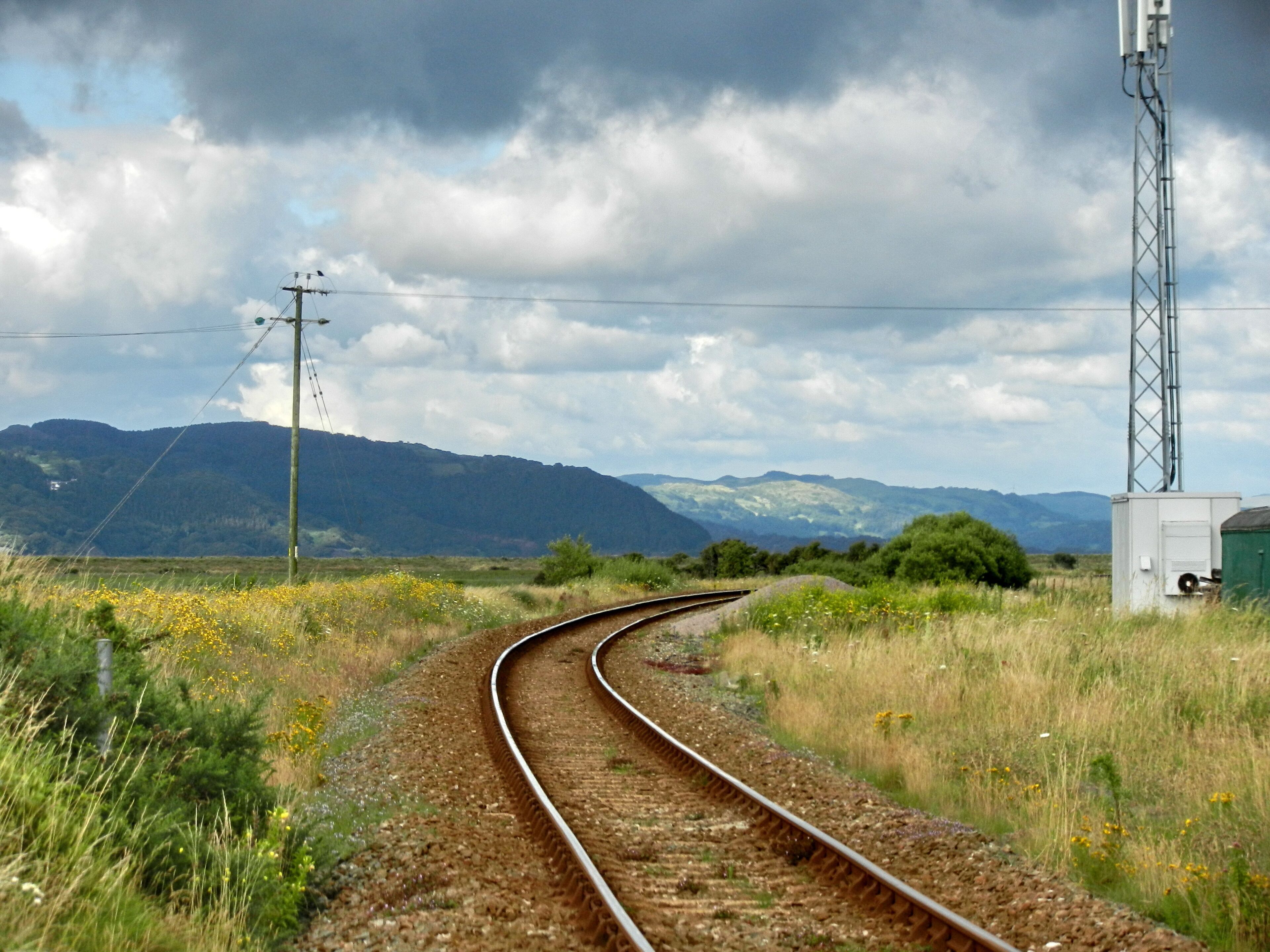 A railway track curving through a grassy field with mountains in the background and a cloudy sky.