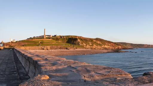 A coastal landscape with a rocky breakwater, a grassy hill with a lighthouse, and the sea.