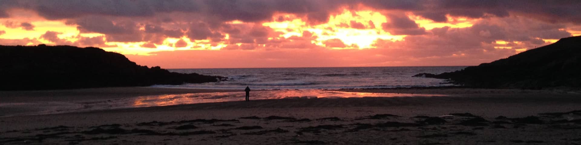 A beach at sunset with dramatic clouds and silhouettes of hills.