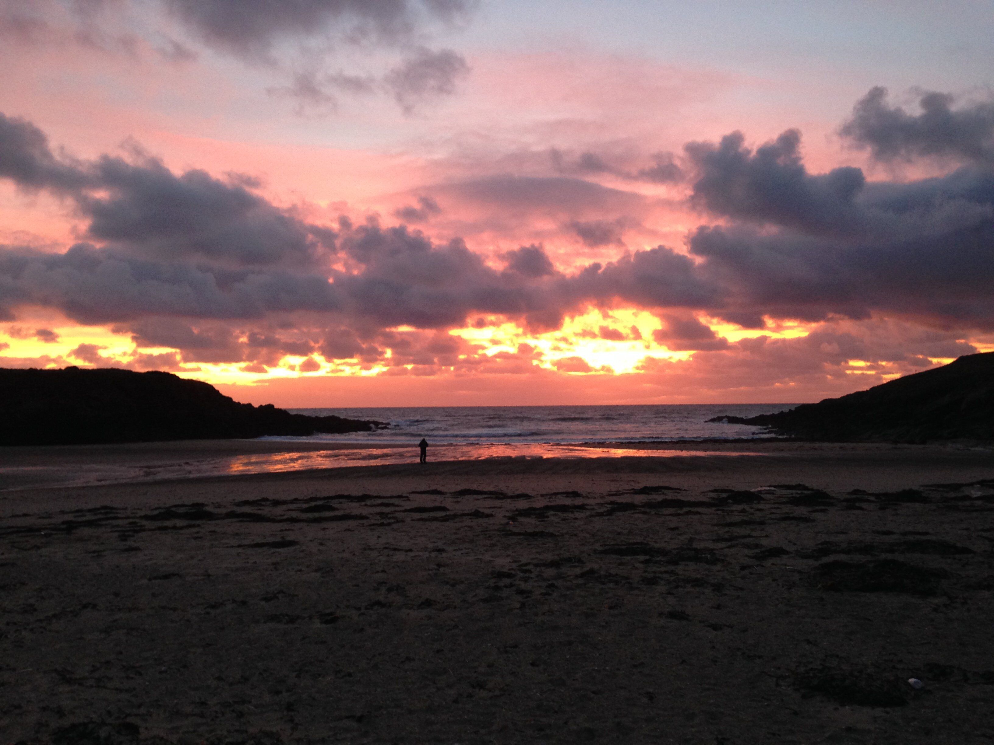 A beach at sunset with dramatic clouds and silhouettes of hills.
