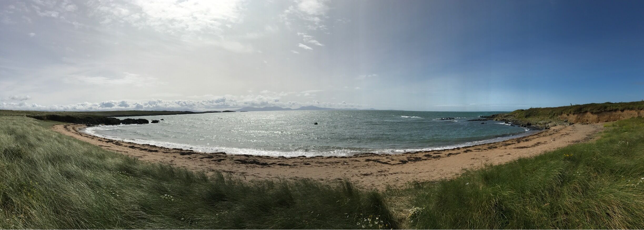 Un paisaje costero con una playa de arena, dunas cubiertas de hierba y un mar tranquilo bajo un cielo parcialmente nublado.