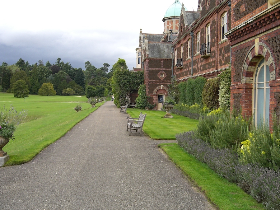 A historic building with a paved pathway leading to a bench, surrounded by well-maintained gardens and trees.