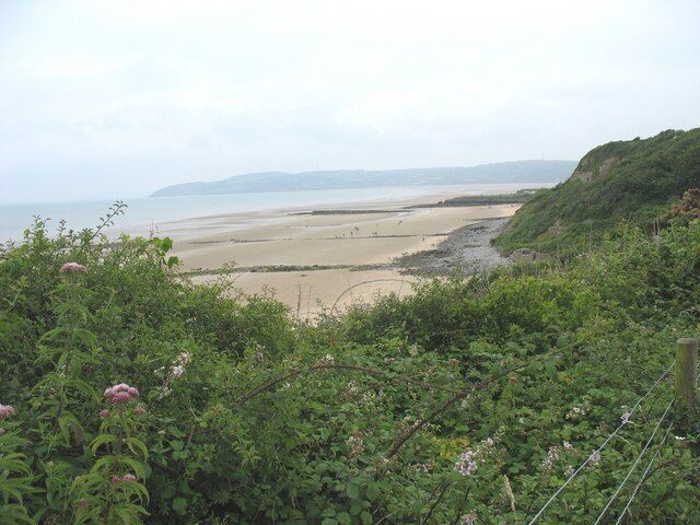 A coastal landscape with a sandy beach, dense greenery, and a distant view of the sea.