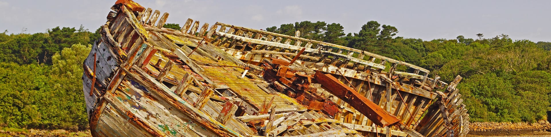 An old, weathered boat stranded on a gravel beach with a forested hillside in the background.