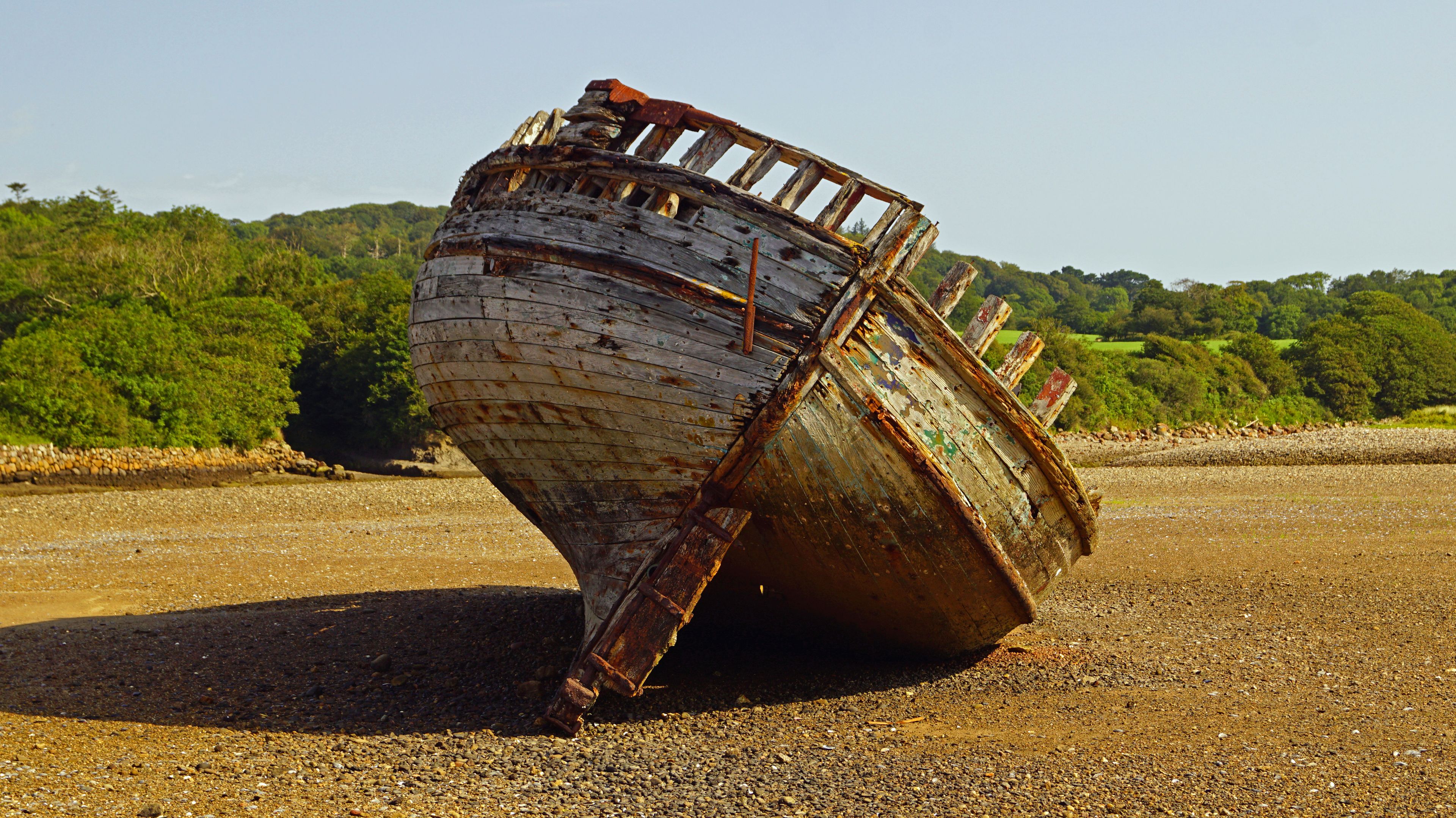 A sunken boat on a gravel beach with a forested hill in the background.