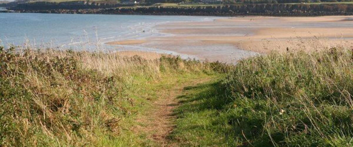 A coastal path leading to a beach with a clear view of the sea and distant hills.