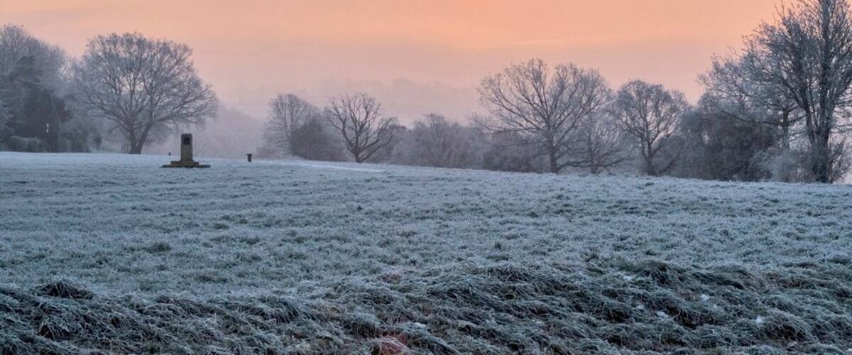 A frost-covered field with a monument in the distance and trees silhouetted against a sunset.