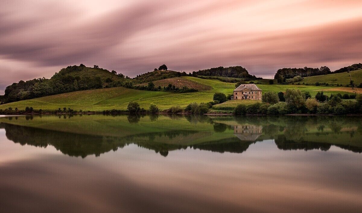 A serene landscape with a house on a hill, a calm lake, and rolling green hills.