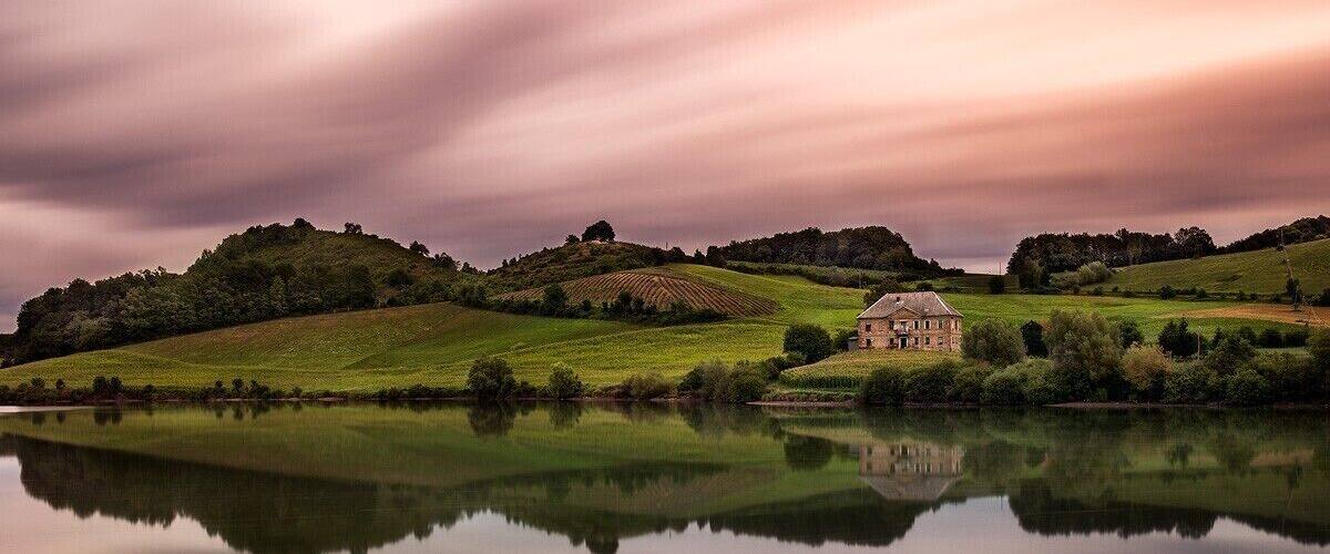 A serene landscape with a house on a hill, a calm lake, and rolling green hills.