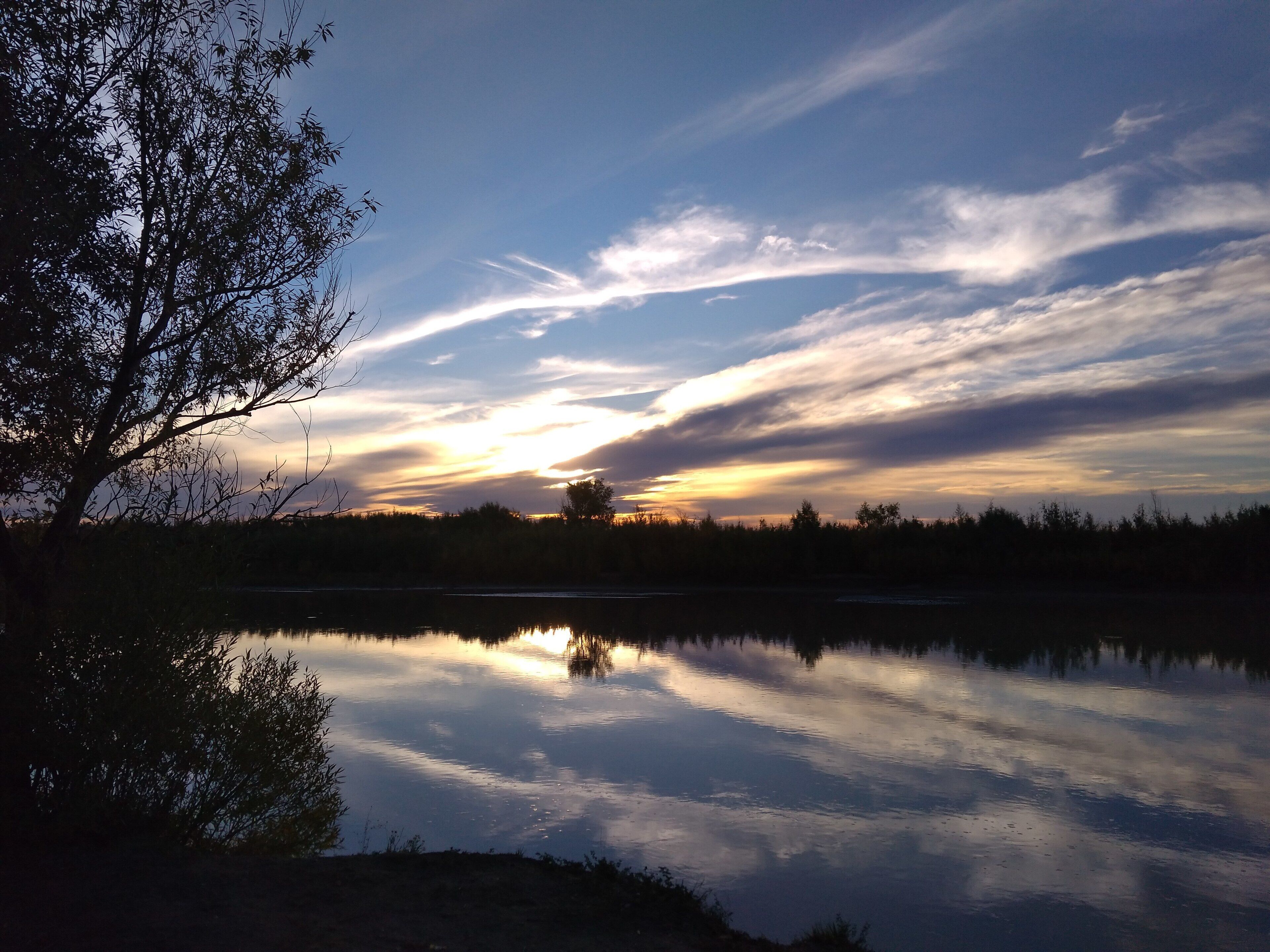 A sunset over a calm lake with reflections of the sky, surrounded by trees and a clear horizon.