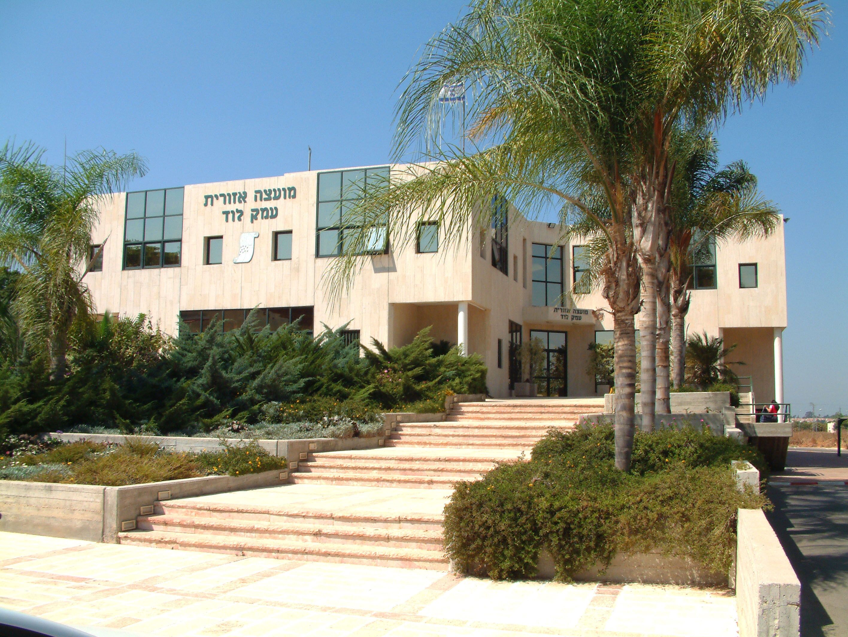 A modern building with Hebrew text on the facade, surrounded by palm trees and a landscaped garden.
