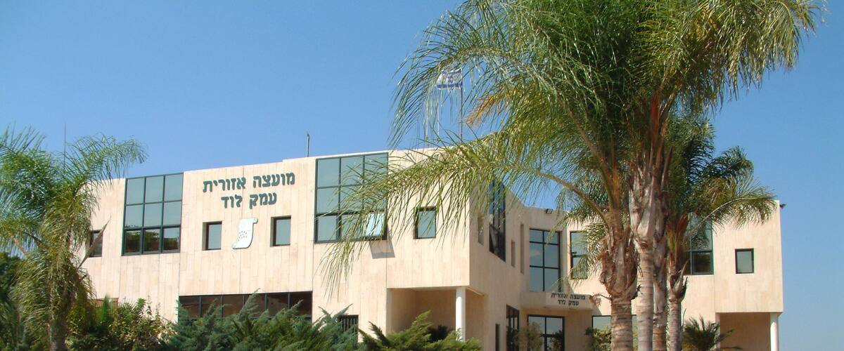 A modern building with Hebrew text on the facade, surrounded by palm trees and a landscaped garden.
