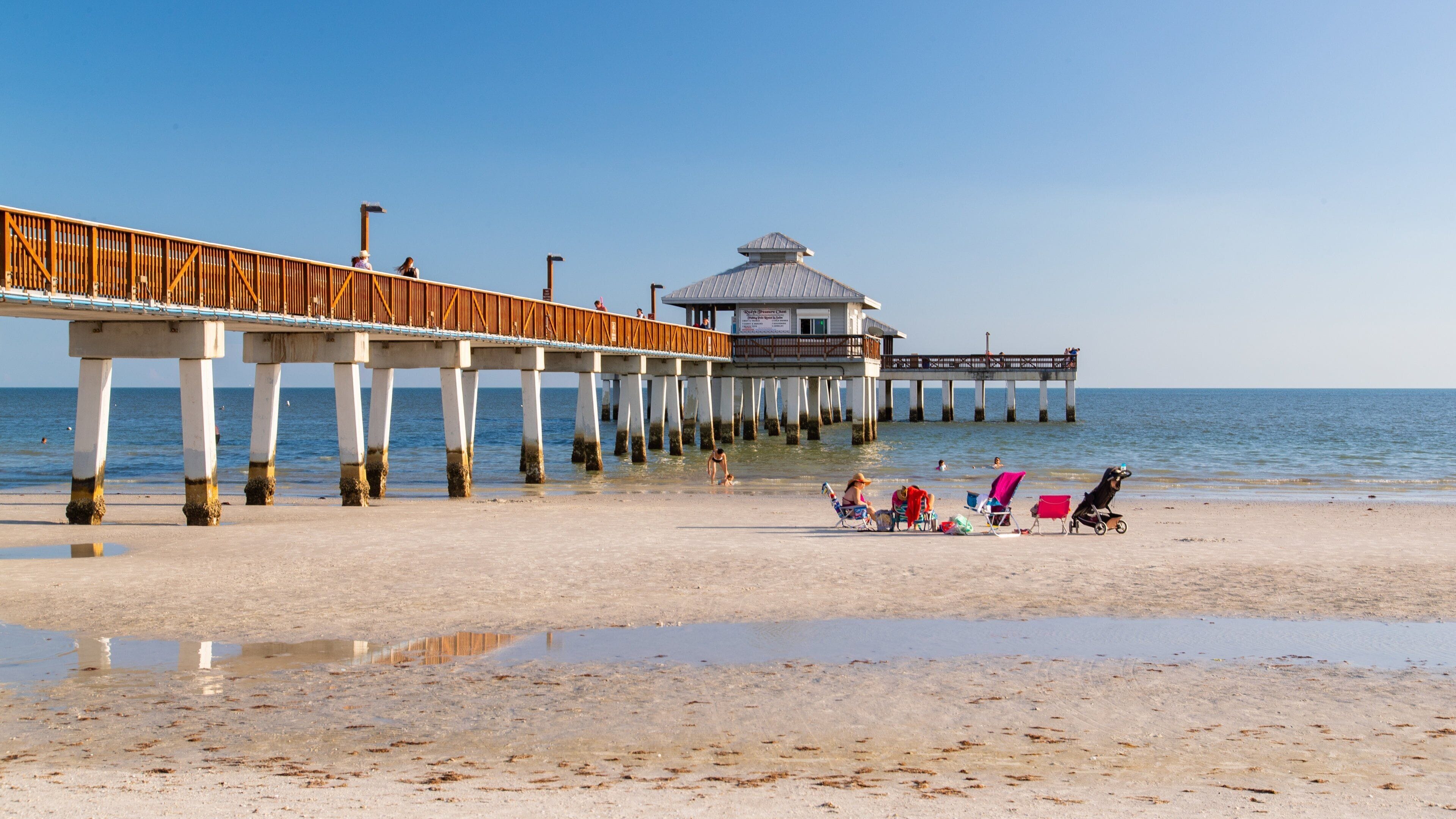 A beach with a wooden pier extending into the ocean, a lifeguard tower, and beachgoers.