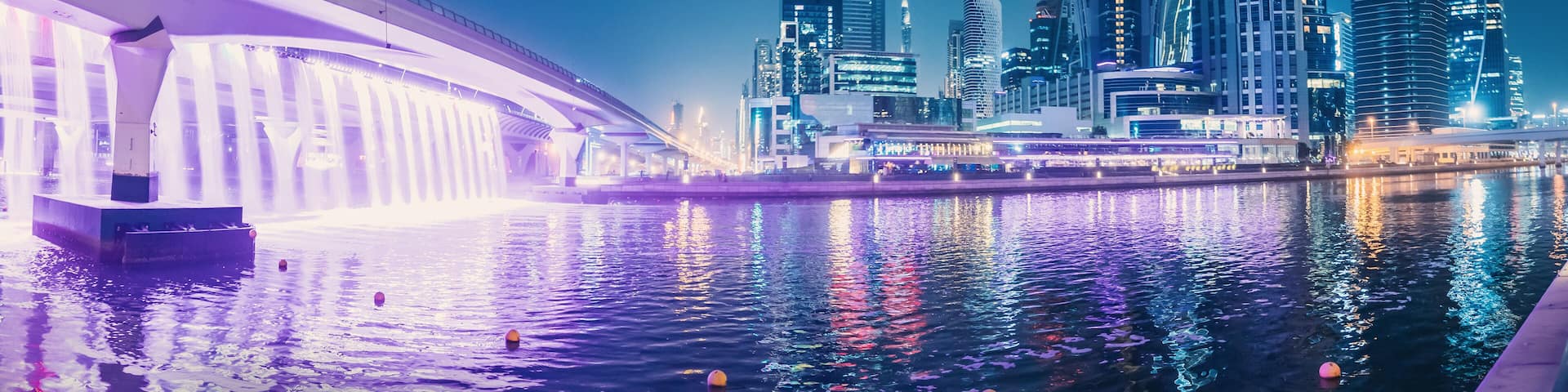 A nighttime cityscape with illuminated skyscrapers and a bridge over a body of water.