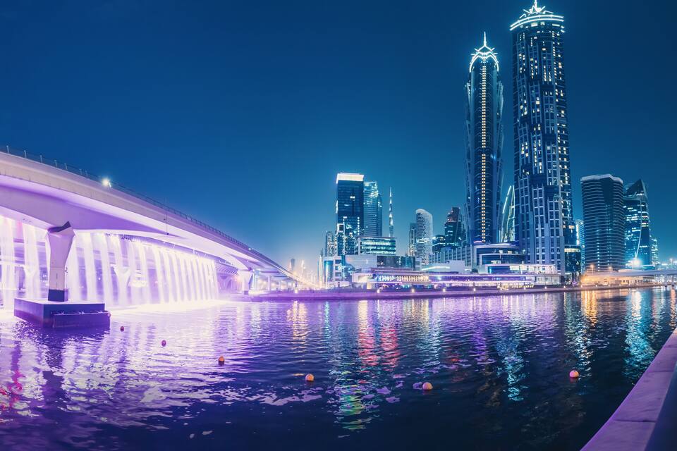 A nighttime cityscape with illuminated skyscrapers and a bridge over a body of water.