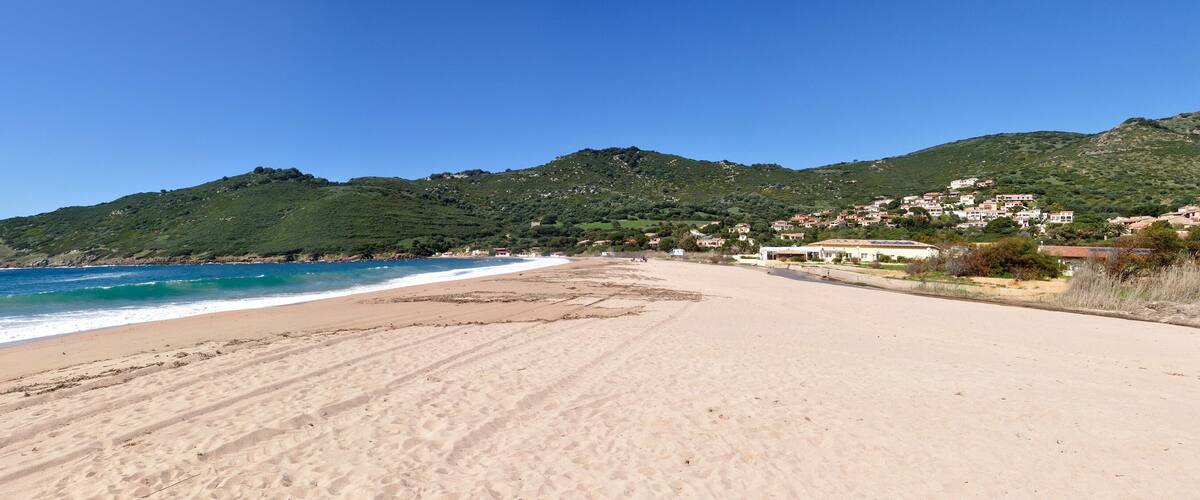 A sandy beach with waves, a clear sky, and a hillside with buildings.