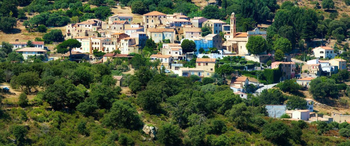 A hillside village with terracotta-roofed buildings, dense greenery, and a prominent church tower.