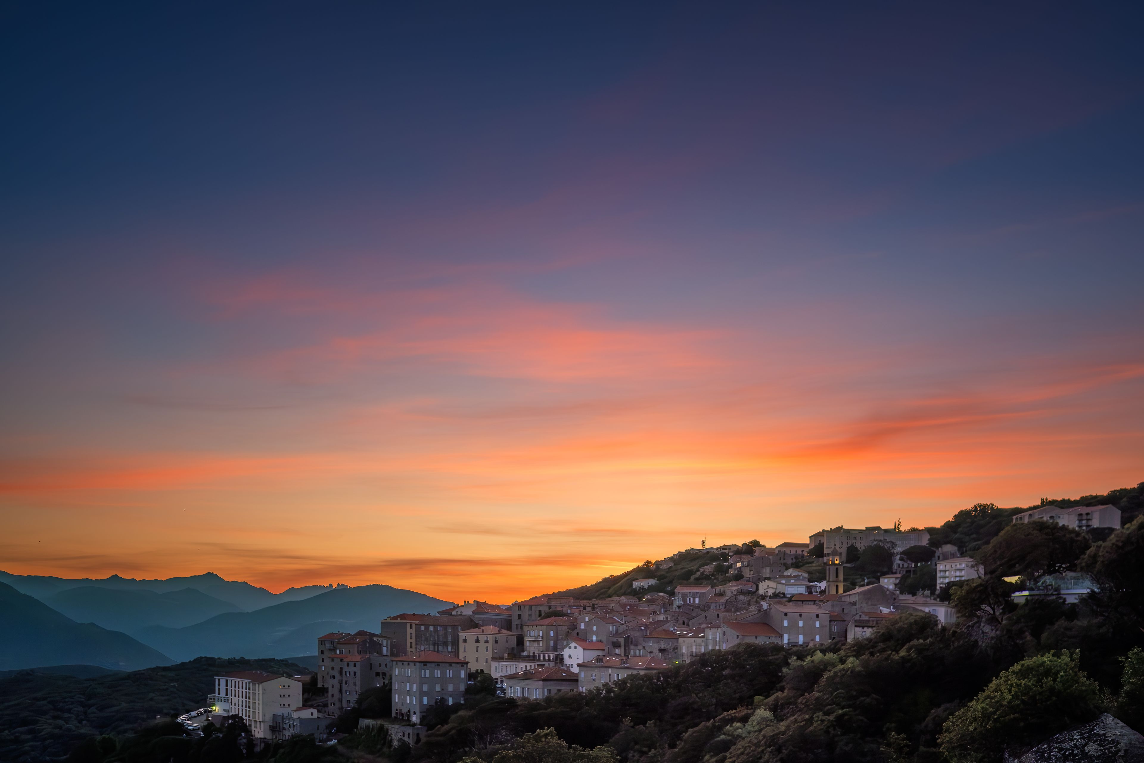 Une ville côtière au coucher du soleil, avec des bâtiments sur une colline et des montagnes en arrière-plan.
