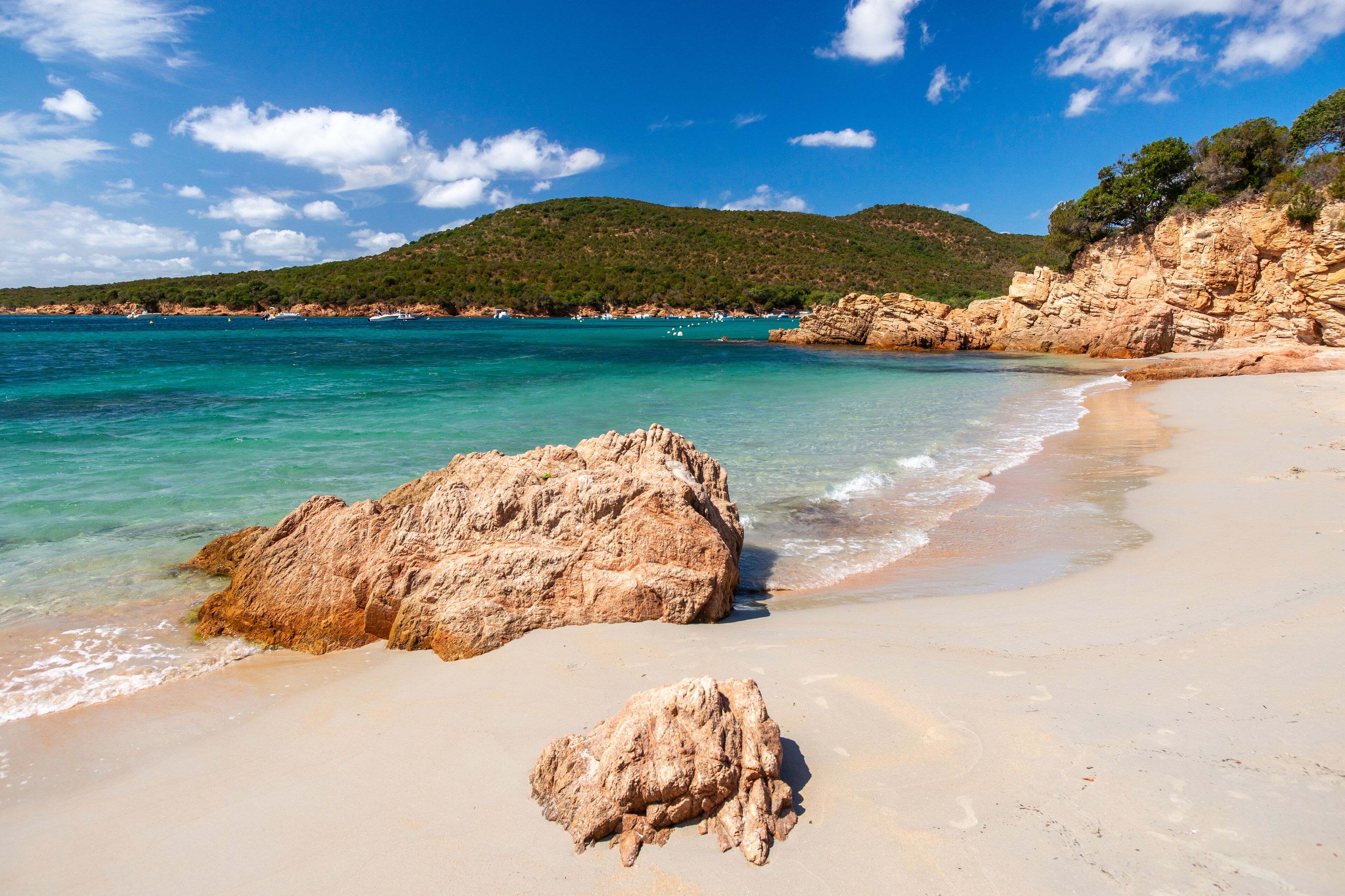 A beach with clear turquoise water, large rocks, and a green hill in the background.