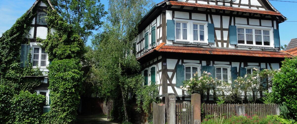 A traditional half-timbered house with a steeply pitched roof, surrounded by lush greenery and a wooden fence.