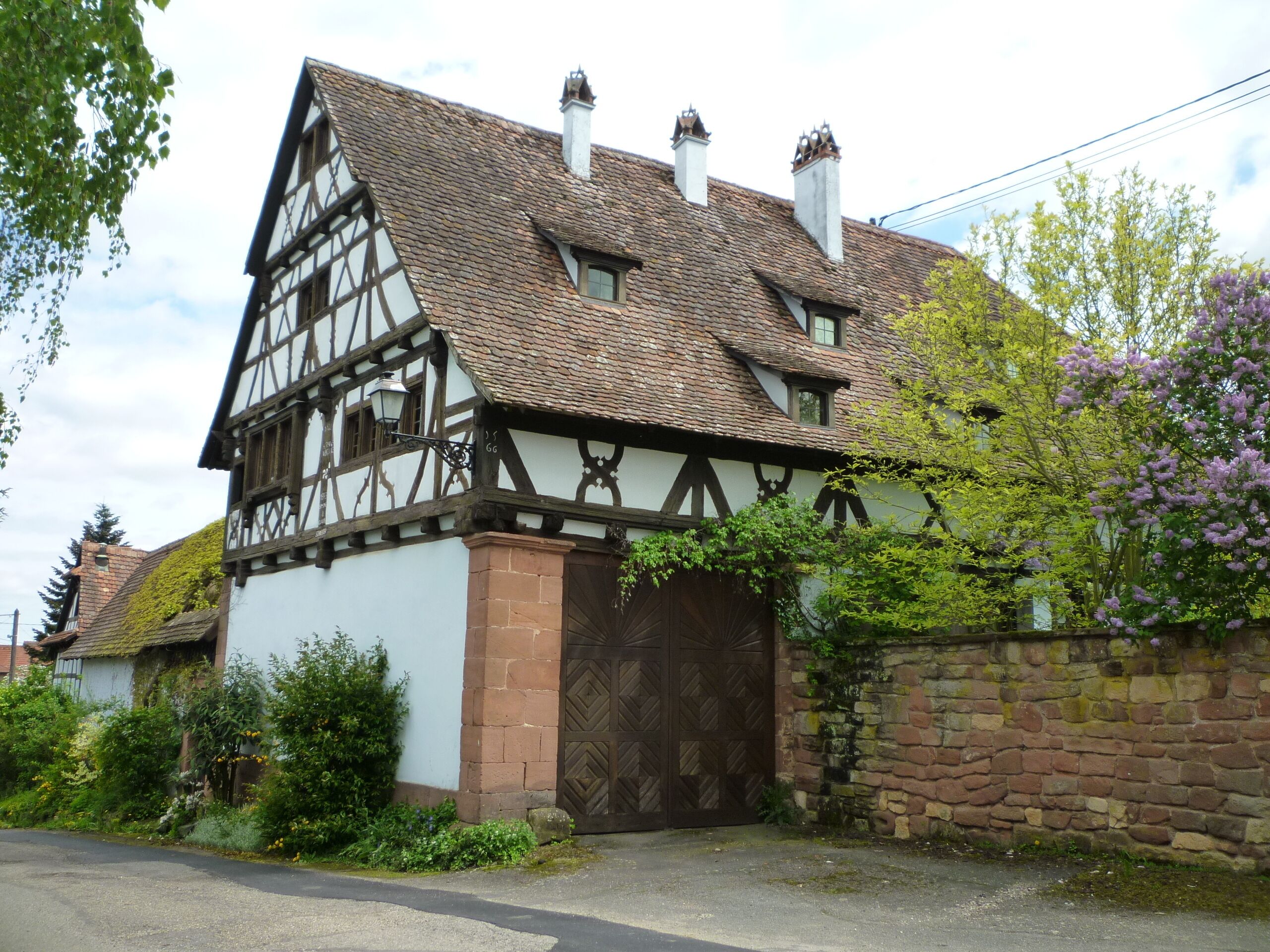 A traditional half-timbered house with a steeply pitched roof, a large wooden door, and a stone wall.