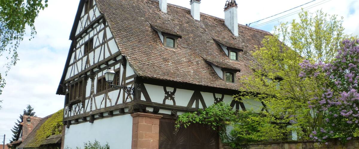 A traditional half-timbered house with a steeply pitched roof, a large wooden door, and a stone wall.