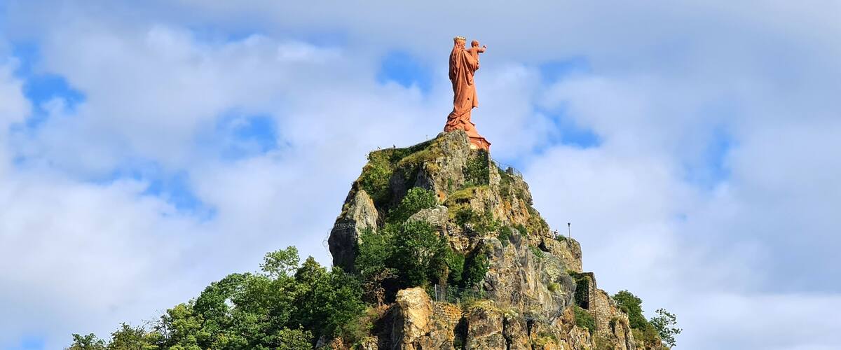 A large statue on a rocky mountain peak with lush greenery at its base.