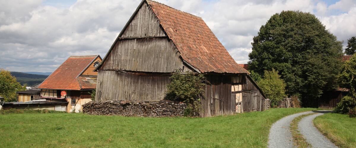 A rustic barn with a sloped roof, surrounded by a gravel path and lush greenery.