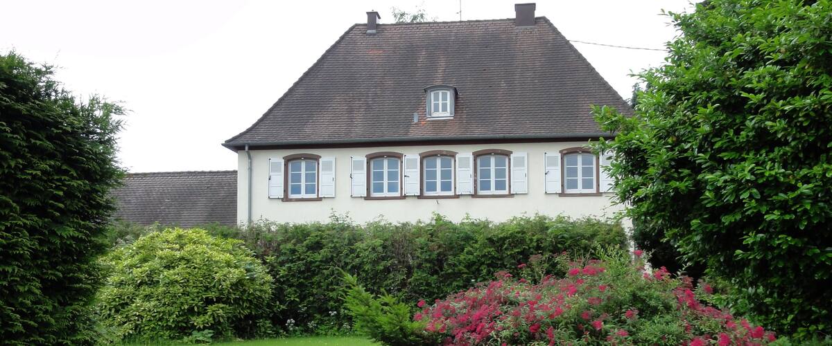 A house with a sloped roof and white shutters, surrounded by a well-maintained lawn and flowering bushes.