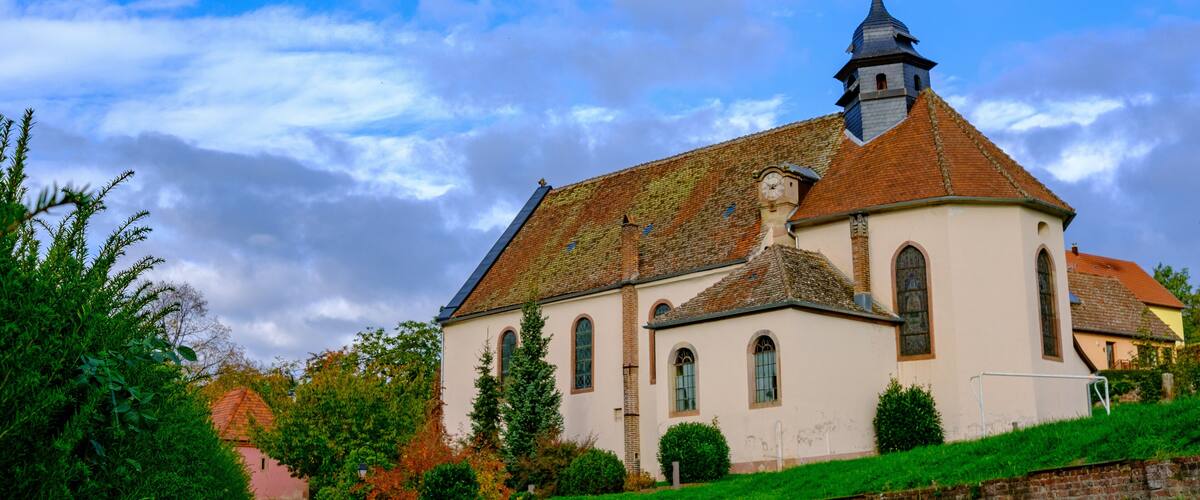 A church with a red roof and a bell tower, surrounded by greenery and under a partly cloudy sky.