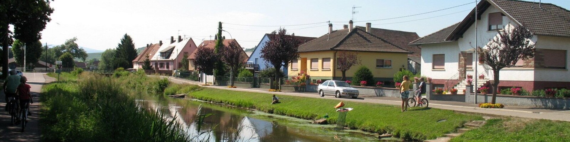 A canal with houses on one side and a road with pedestrians and cyclists on the other.