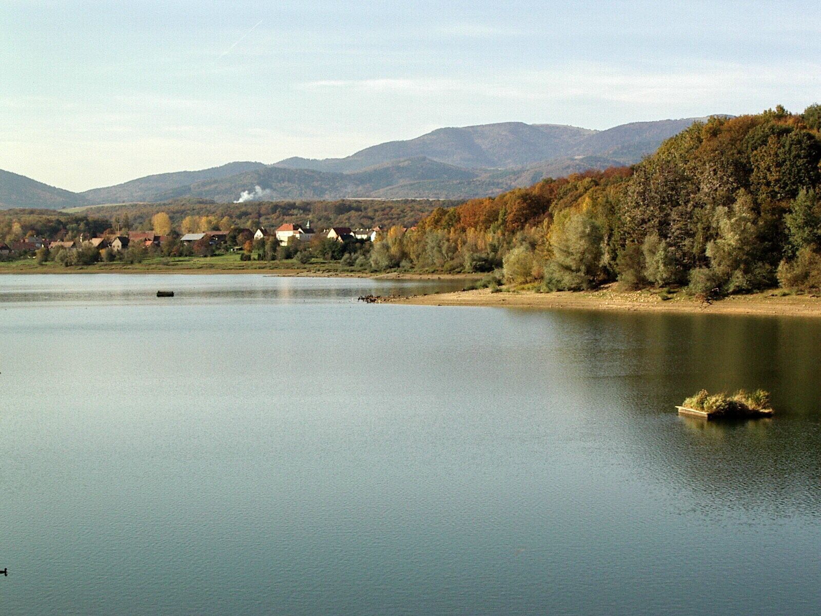 A calm lake with a small boat, surrounded by houses and autumn-colored trees, with mountains in the background.