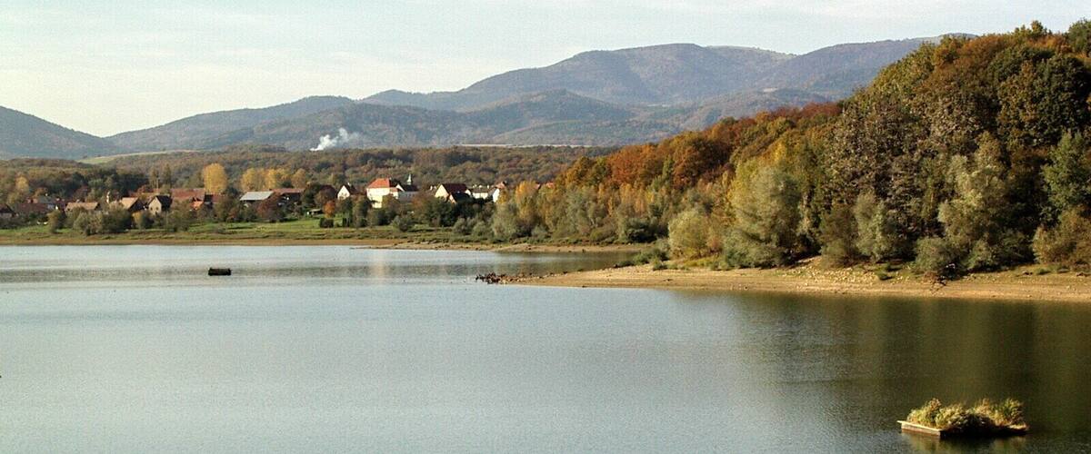 A calm lake with a small boat, surrounded by houses and autumn-colored trees, with mountains in the background.