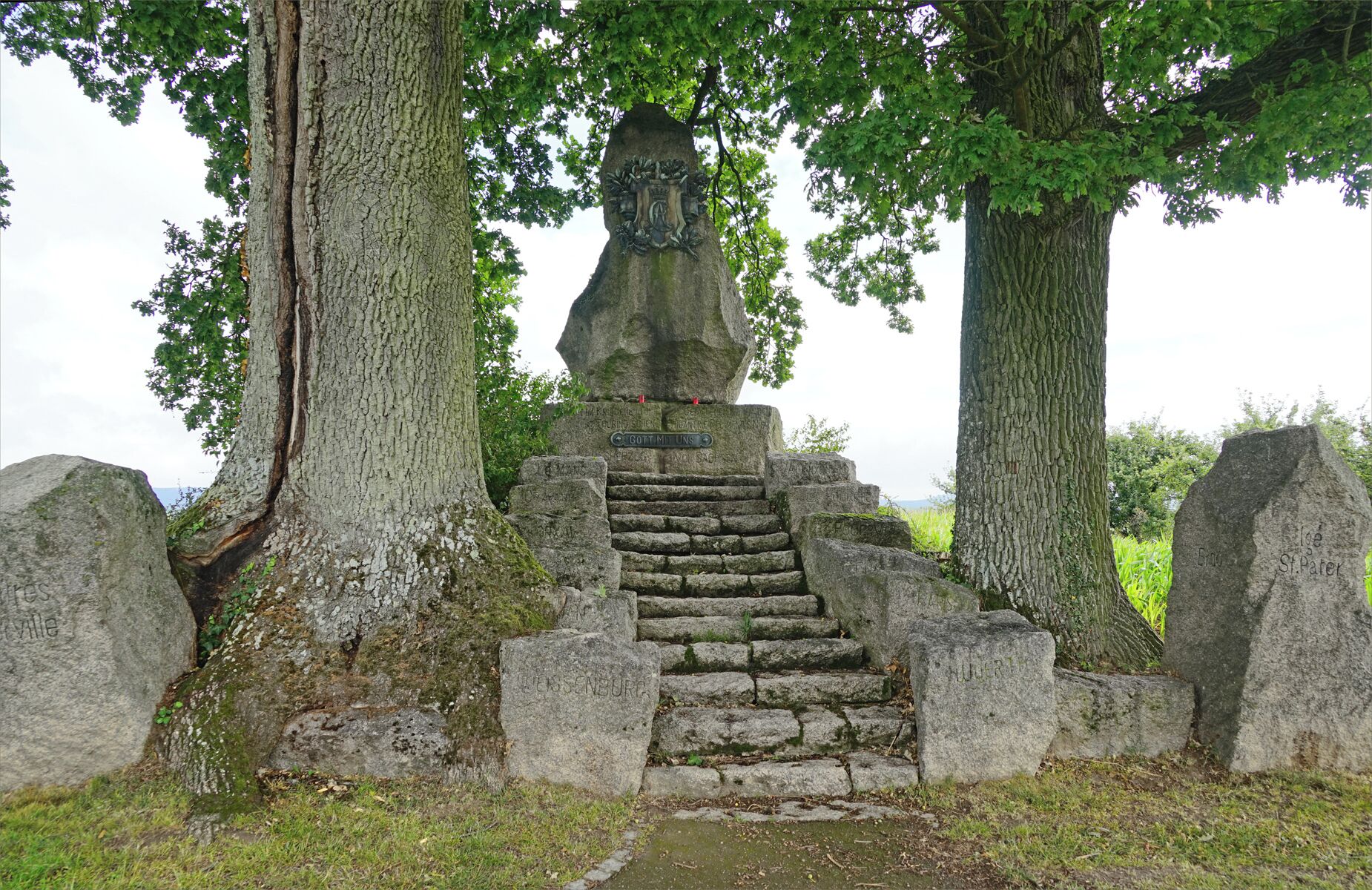 A stone monument with inscriptions and a carved coat of arms, surrounded by large trees and a grassy area.
