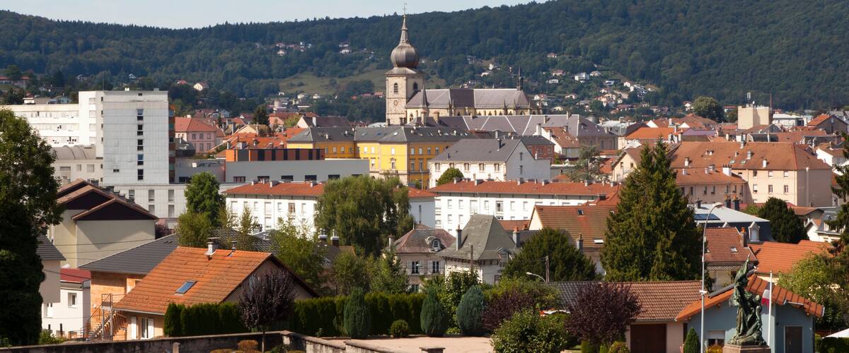 A town with a prominent church tower, surrounded by residential buildings and a forested hill in the background.