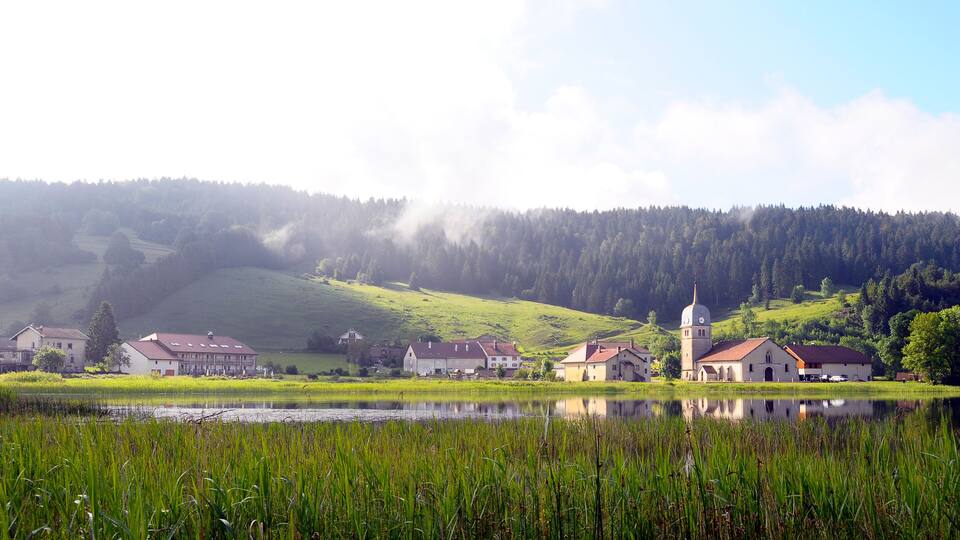 A serene lakeside village with houses and a church surrounded by lush greenery and hills.