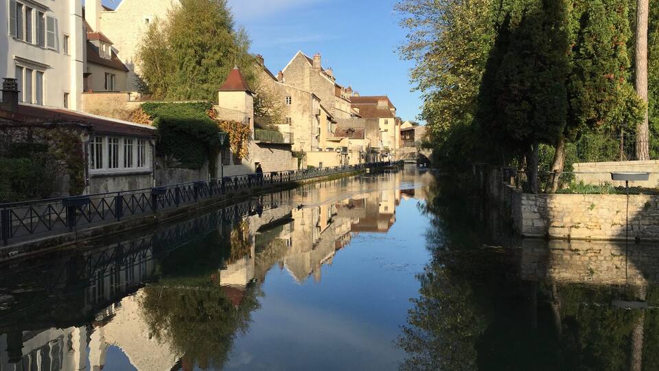A calm canal with buildings reflecting on the water, surrounded by trees and stone walls.