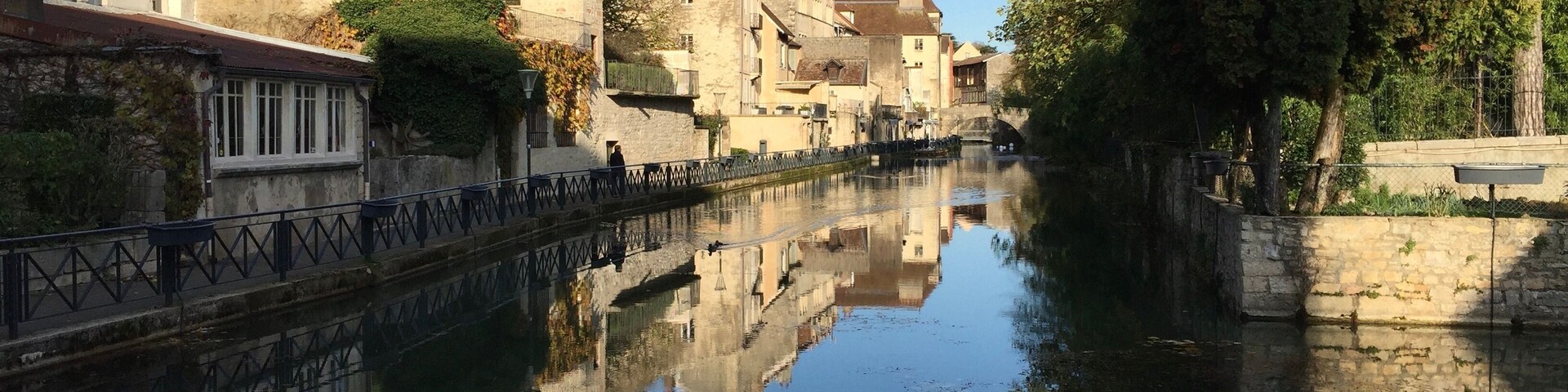 Un canal tranquilo con edificios reflejándose en el agua, rodeado de árboles y muros de piedra.