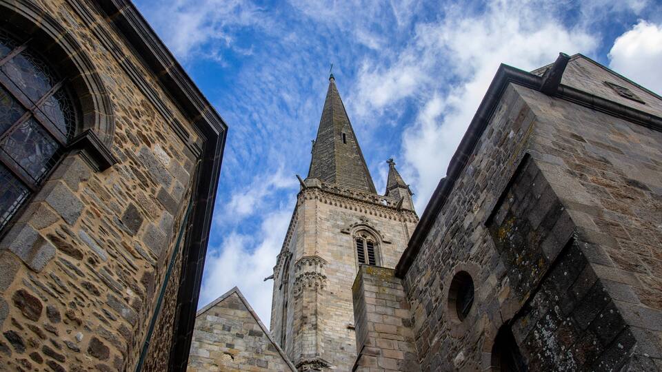 A historic stone tower with a pointed spire and arched windows, surrounded by other stone buildings.