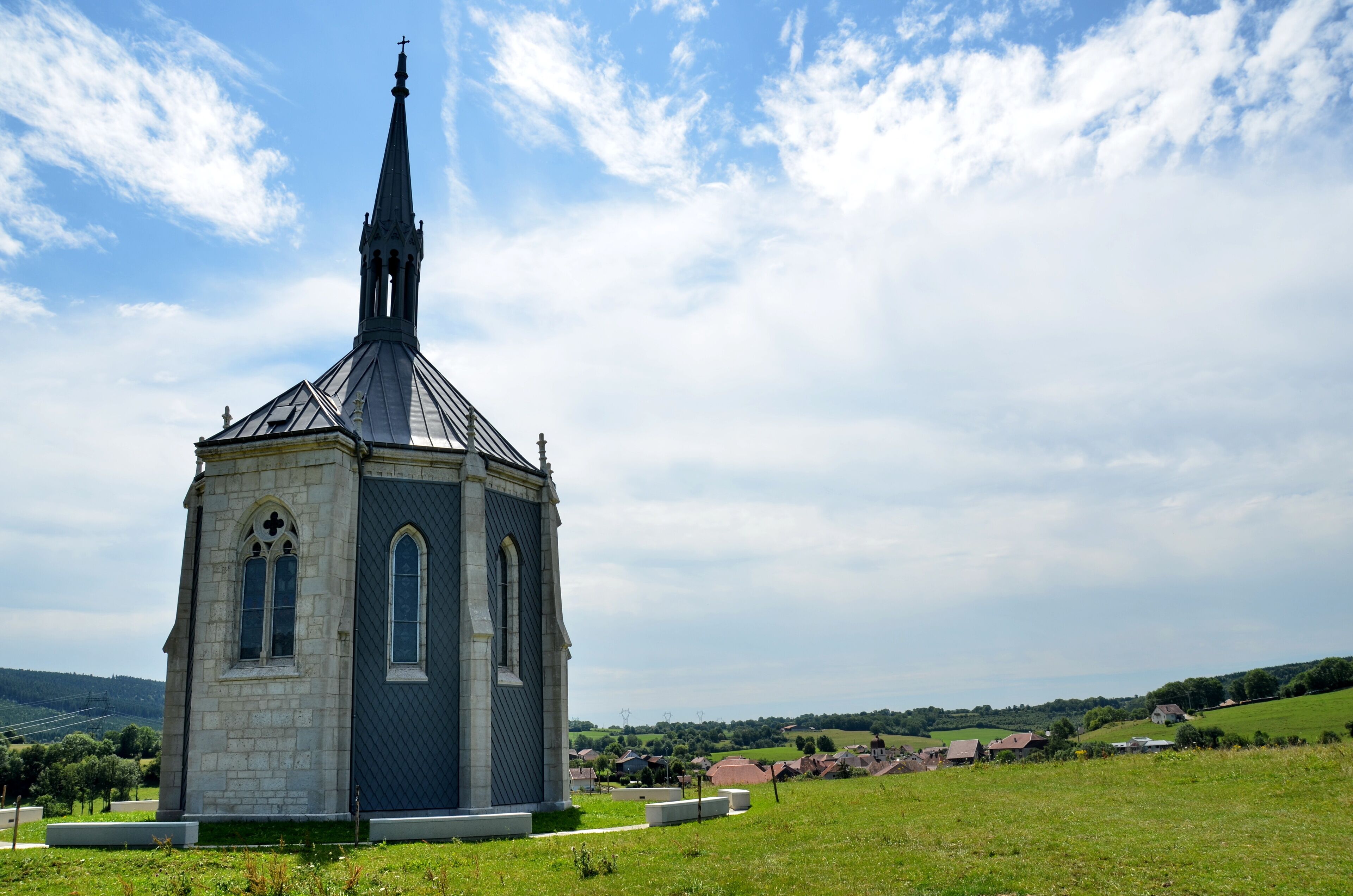 A church with a tall steeple and a small tower, surrounded by a green field and a village in the background.