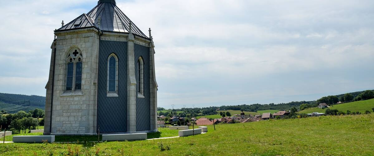 A church with a tall steeple and a small tower, surrounded by a green field and a village in the background.