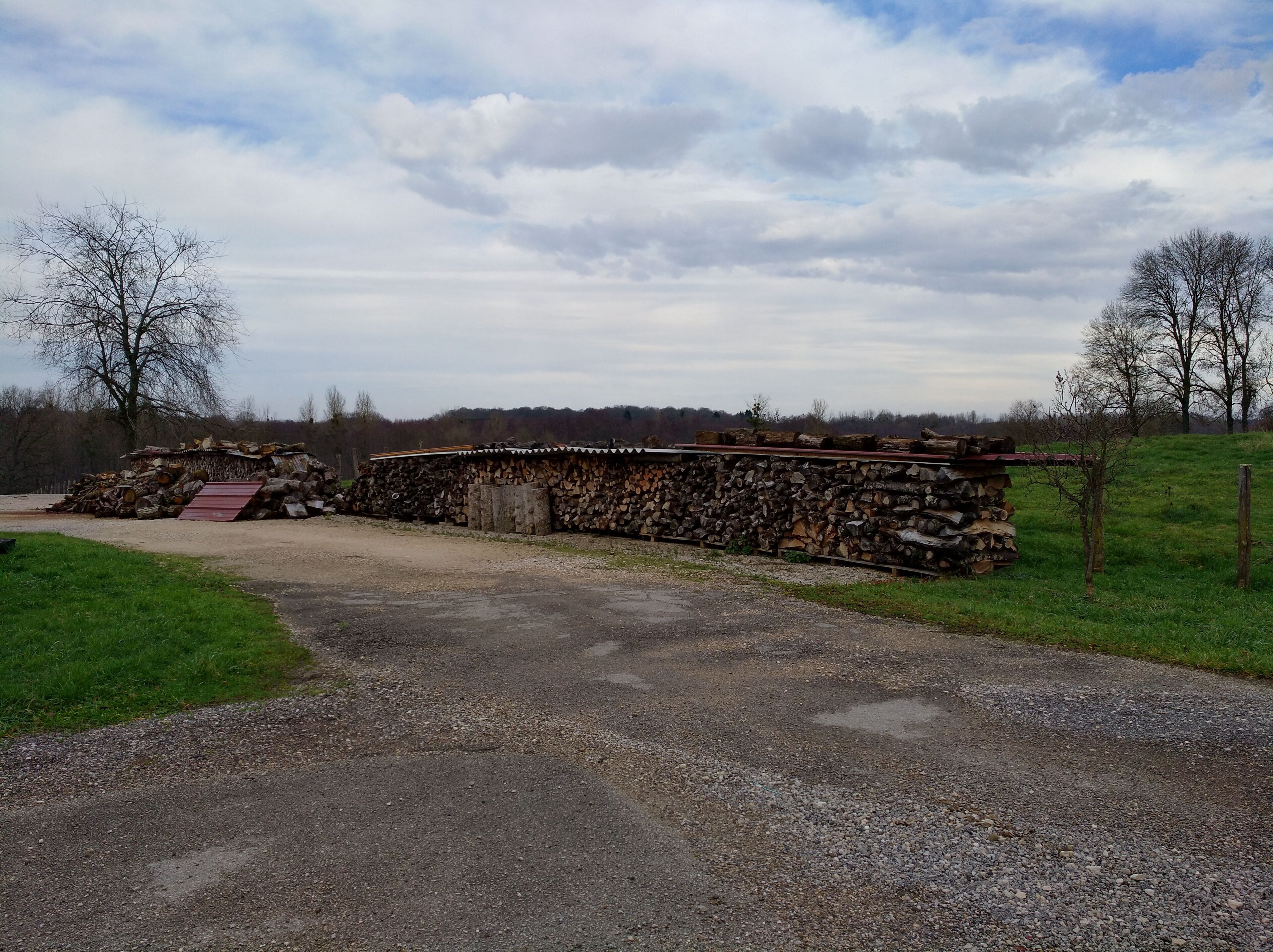 A gravel road leading to a stone wall with stacked firewood.