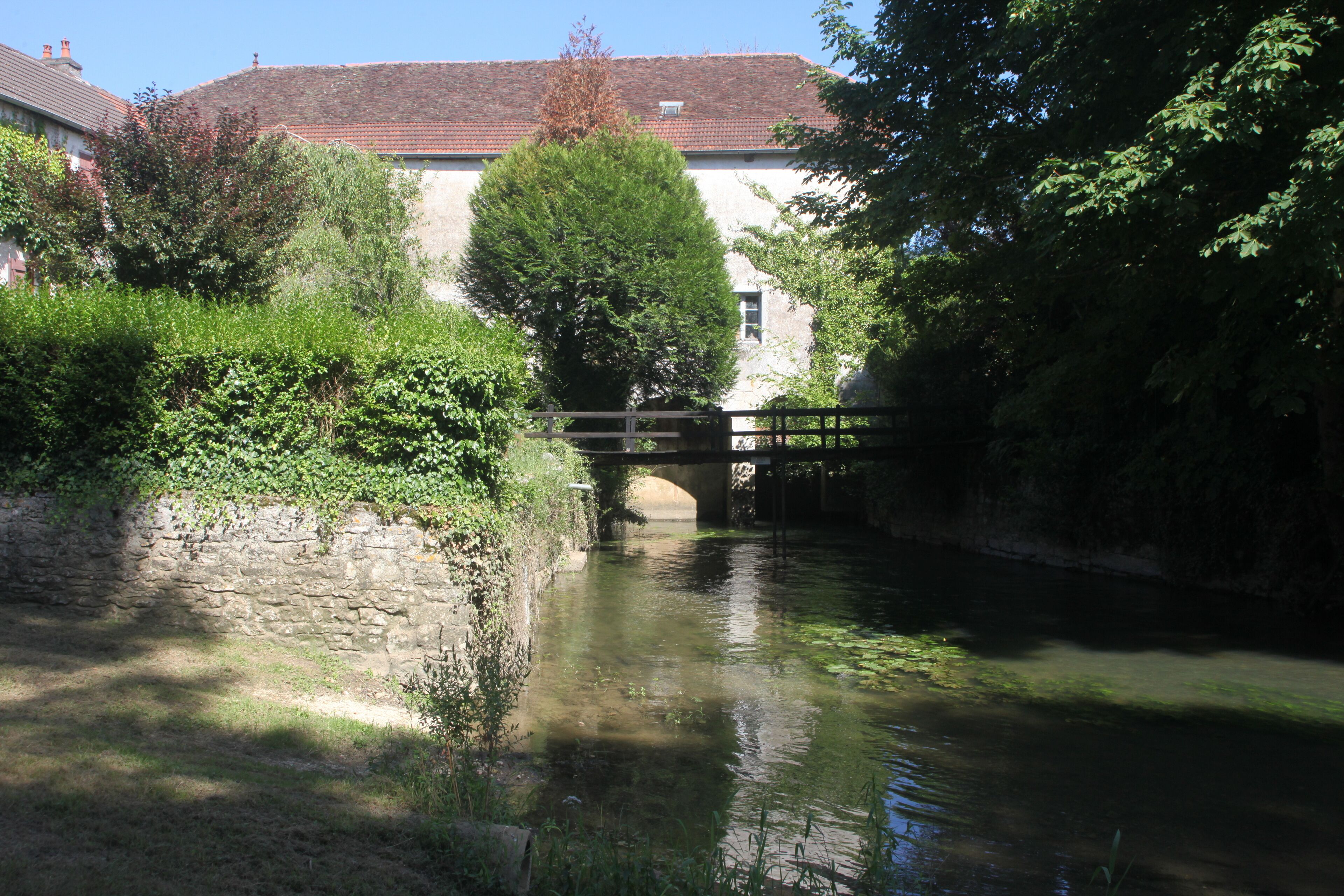 A stone bridge over a calm river, surrounded by lush greenery and a building with a tiled roof.