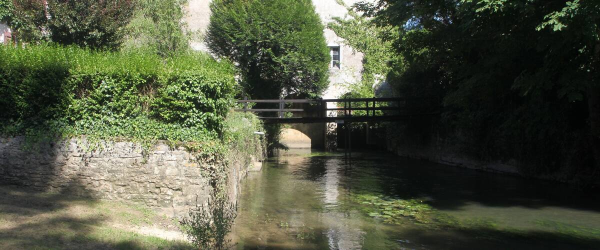 A stone bridge over a calm river, surrounded by lush greenery and a building with a tiled roof.