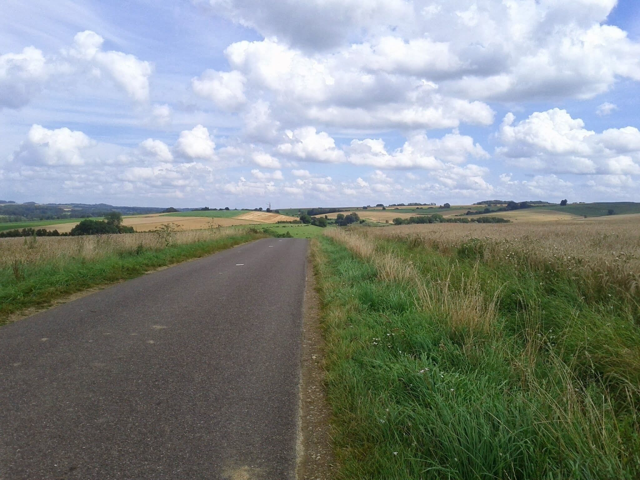 A rural road flanked by fields and rolling hills under a partly cloudy sky.