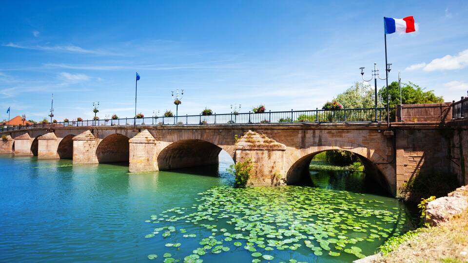 A stone bridge over a river with lily pads and a French flag.