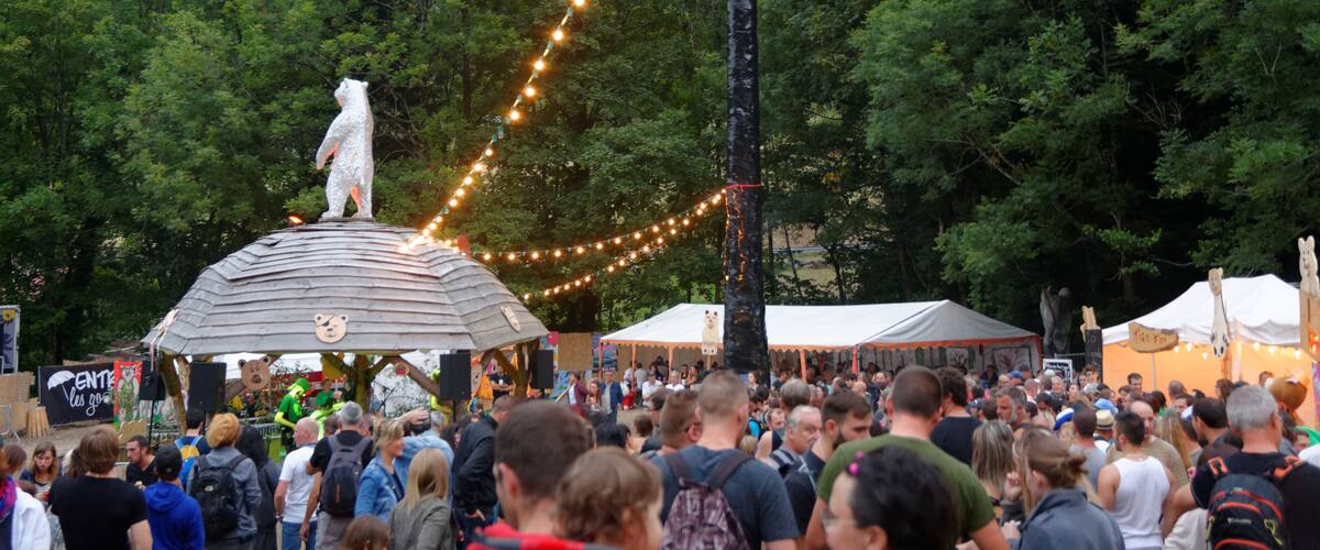 A crowd at a festival with a bear statue on a structure, surrounded by trees and string lights.